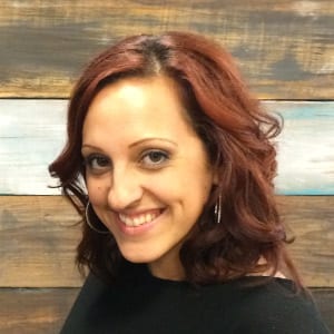 A stylist with dark red wavy hair and hoop earrings, smiling in front of the salon’s painted plank wall.