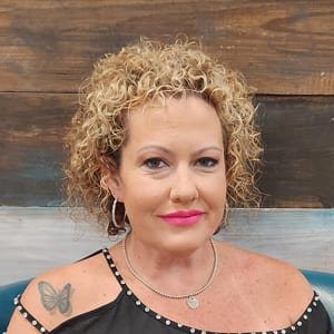 A stylist with short curly blonde hair, smiling in front of the salon’s painted plank wall.