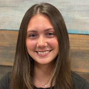 A stylist with long straight brown hair, smiling in front of the salon’s painted plank wall.