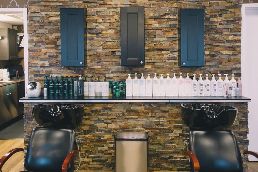 Two black shampoo bowls under a stacked stone wall, with a long shelf of shampoo and conditioner bottles above them.